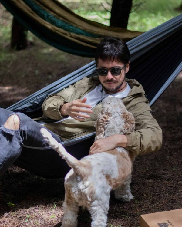 Foto de un hombre acariciando un perro al borde de una Hamaca en el bosque